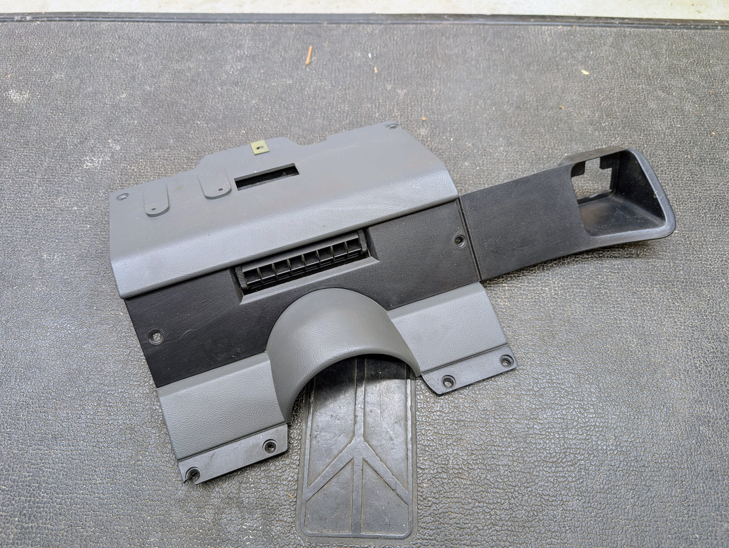 A General Motors OEM Lower Dash Trim Knee Bolster in dark gray for 1986–1993 Chevy S10, Blazer, GMC S15, and more (part #15591143) with vent, cutout, and screw holes rests on a textured mat.