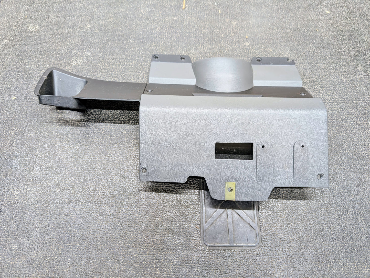 A General Motors OEM Lower Dash Trim Knee Bolster in dark gray for 1986-1993 Chevy S10, Blazer, and GMC S15 with a round protrusion, angled duct, and cutouts lies on a textured dark surface.