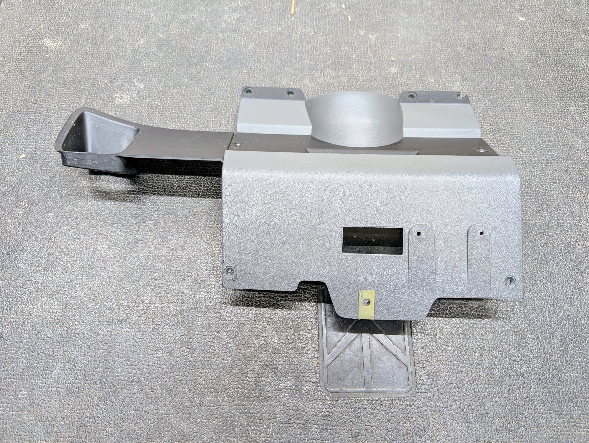 A General Motors OEM Lower Dash Trim Knee Bolster in dark gray for 1986-1993 Chevy S10, Blazer, and GMC S15 with a round protrusion, angled duct, and cutouts lies on a textured dark surface.