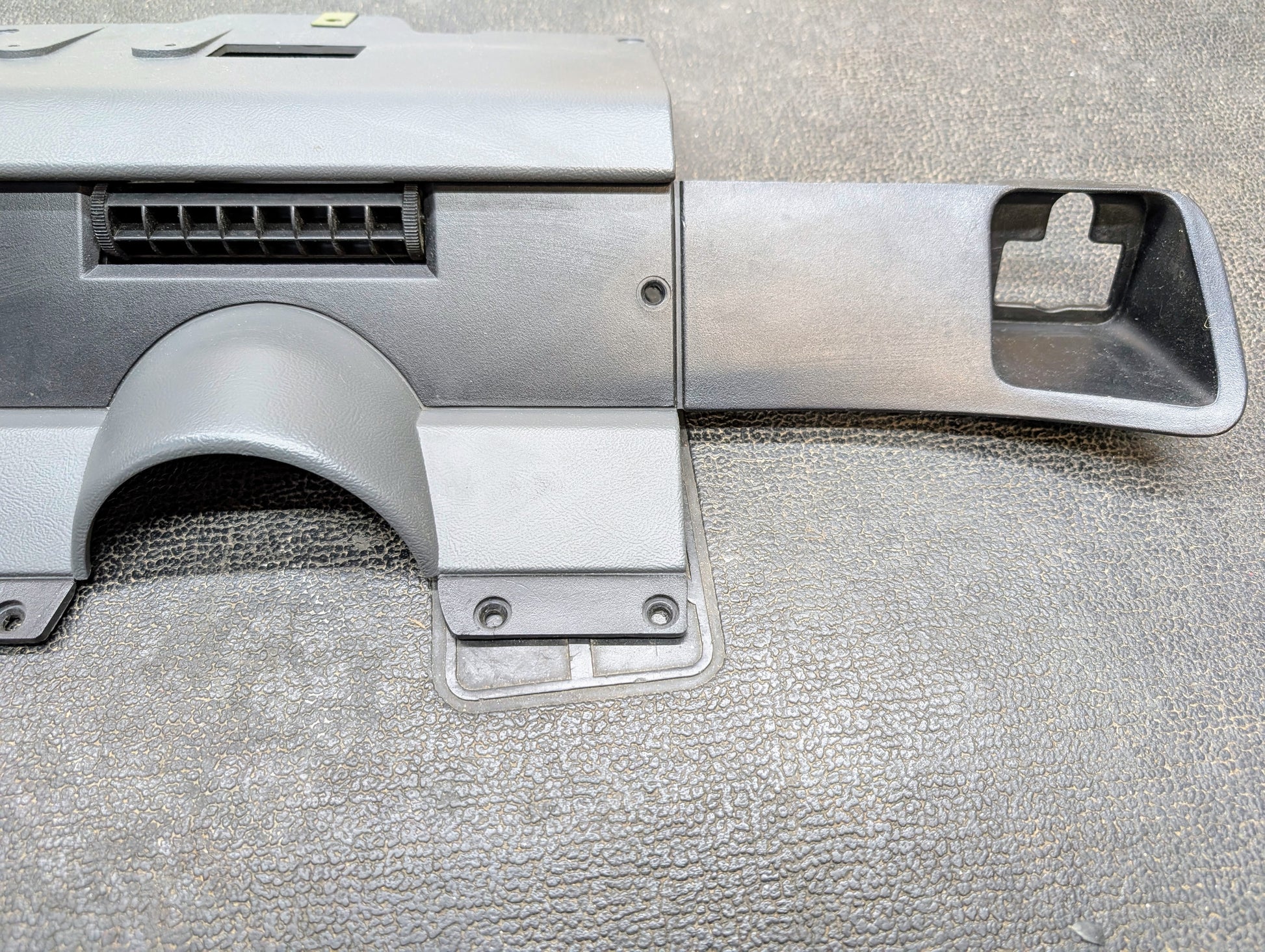 A close-up of a General Motors OEM Lower Dash Trim Knee Bolster in Dark Gray for 1986-1993 Chevy S10, Blazer, GMC S15, and more is shown with an air vent and rectangular cutout, laid on a textured surface.