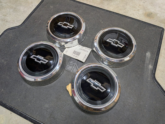 Four NOS Bowtie Dog Dish 10.5" hub caps by General Motors for 1982–1999 Chevy S10, Blazer, Astro, and more are arranged on a black mat with a white label showing—these genuine GM parts feature black-and-silver Chevy logos.