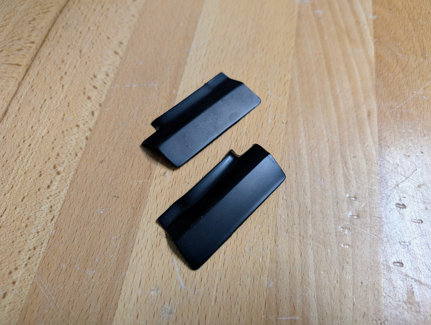 Two small, black plastic trim pieces, likely NOS rear window molding panel covers for 1991–1994 Chevy S10, Blazer, GMC S15 Jimmy (General Motors), are shown on a light wood surface; each has grooves and angled edges.
