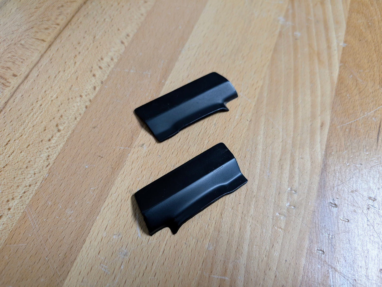 A pair of NOS rear window molding trim panel covers by General Motors for 1991–1994 Chevy S10, Blazer, GMC S15 Jimmy, and more are shown parallel on a light wooden surface. The small black plastic pieces have curved edges.