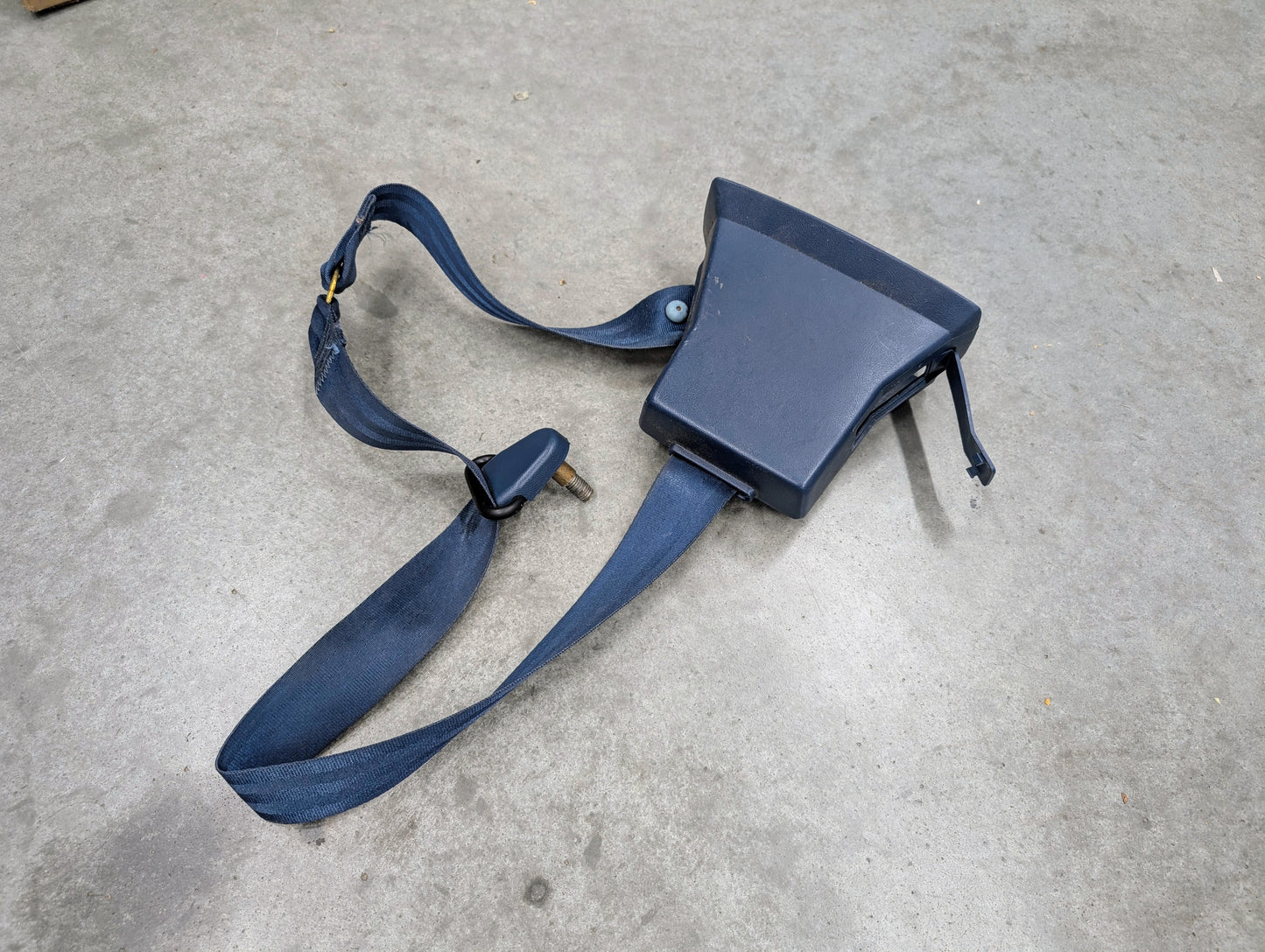 A General Motors OEM driver seat belt retractor and buckle in blue for 1982–1993 Chevy S10 or GMC S15 rests on a gray concrete surface.