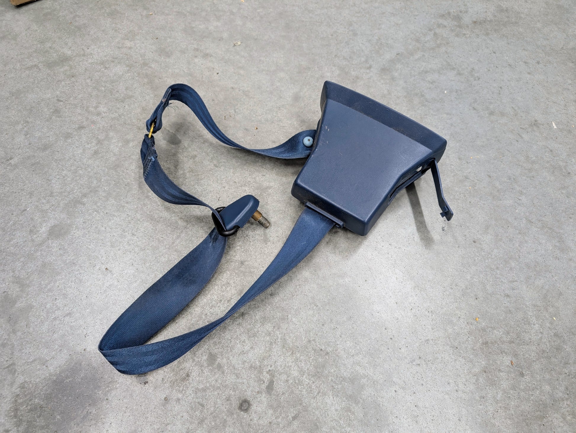 A General Motors OEM driver seat belt retractor and buckle in blue for 1982–1993 Chevy S10 or GMC S15 rests on a gray concrete surface.