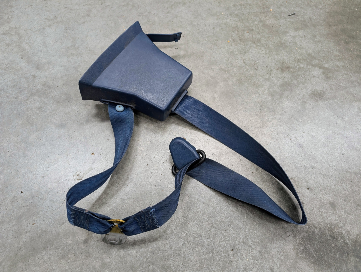 A General Motors OEM driver seat belt retractor and buckle in blue for 1982–1993 Chevy S10 and GMC S15 is lying on a gray concrete surface.