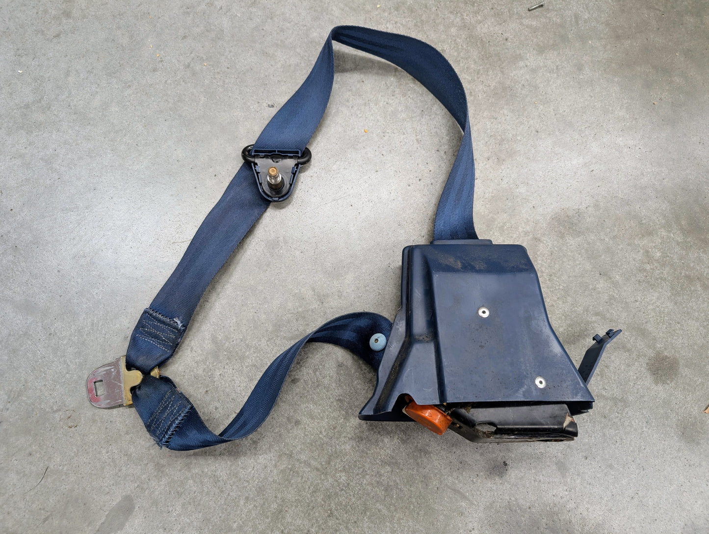 A General Motors OEM blue driver seat belt retractor and buckle for 1982-1993 Chevy S10/GMC S15 is shown partially extended on a gray concrete surface, displaying signs of use.