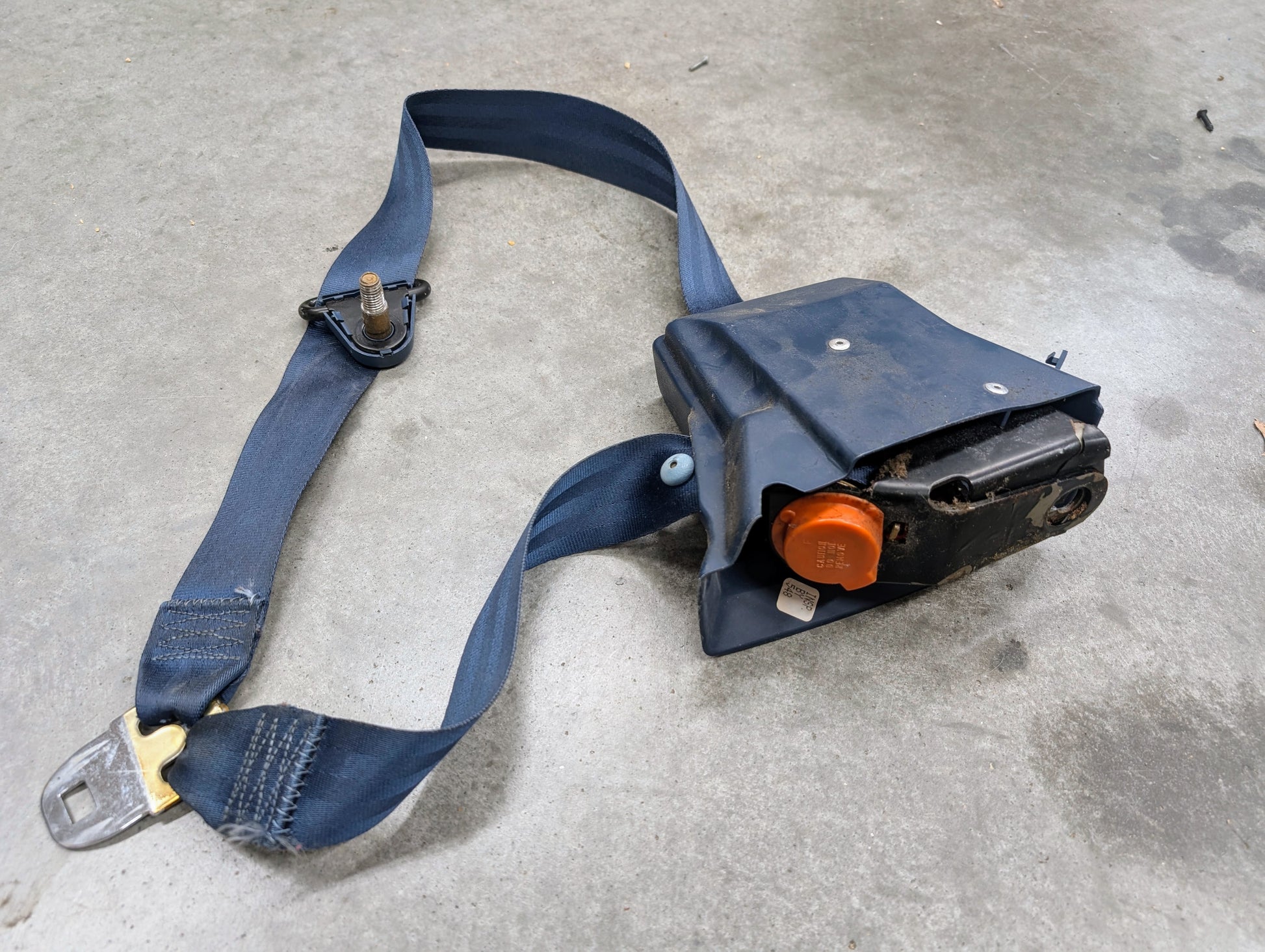 A General Motors OEM blue driver seat belt retractor and buckle for 1982-1993 Chevy S10/GMC S15 is shown on a gray concrete floor. The retractor features an orange button and fits Chevrolet S10 models.